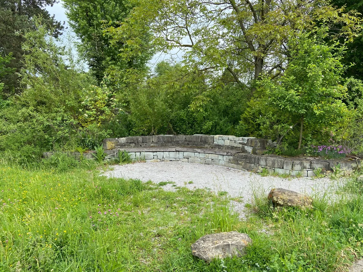 Eine kleine Mauer an einem Platz, davor Wiese und viel Grün.